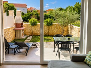 Eine Terrasse mit Grill, Gartenmöbeln und Blick auf eine Steinsäule sowie Grünpflanzen.