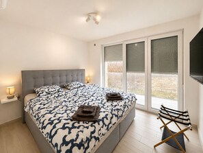 Ein Schlafzimmer mit einem großen Bett, einem Nachttisch mit Lampe, einem Fenster mit Jalousien und einem Klappstuhl.