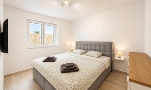 A bedroom with a bed, two bedside lamps, a TV mounted on the wall, and a window with a view of a stone wall. - Apartment Mariner 2 with heated pool (Zaton)