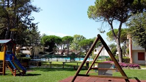 A playground with swings and a slide, surrounded by grass and trees.