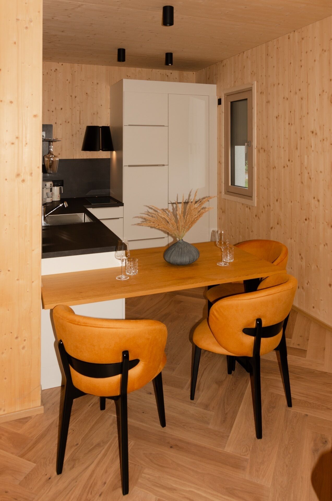 Une cuisine moderne avec des meubles blancs, une cuisinière noire et une table à manger en bois, accompagnée de deux chaises orange.