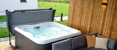 Un jacuzzi con terraza de madera, un sofá gris con cojines y una pared de madera con una lámpara.