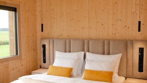 A bedroom with a wooden paneled wall, a large bed with white and yellow bedding, and a window providing a view of the outdoors. - Oaza Luxury Resort (Moravske Toplice)