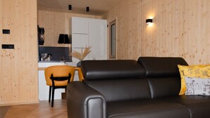 A modern living room with a black leather sofa, a wooden dining table, and a kitchen area with a white cabinet and a small plant. - Oaza Luxury Resort (Moravske Toplice)
