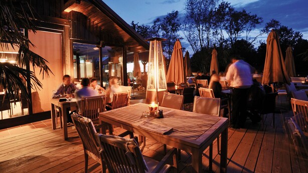 An outdoor dining area with wooden tables and chairs, a central table with a candle, and umbrellas. - Oaza Luxury Resort (Moravske Toplice)