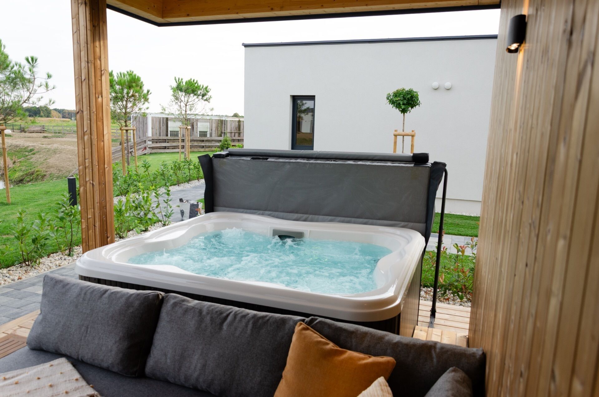 Un jacuzzi con un cojín gris, una terraza de madera y un edificio blanco al fondo.