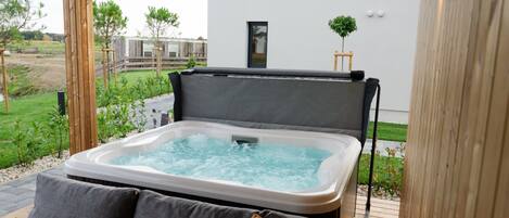 Un jacuzzi con un cojín gris, una terraza de madera y un edificio blanco al fondo.
