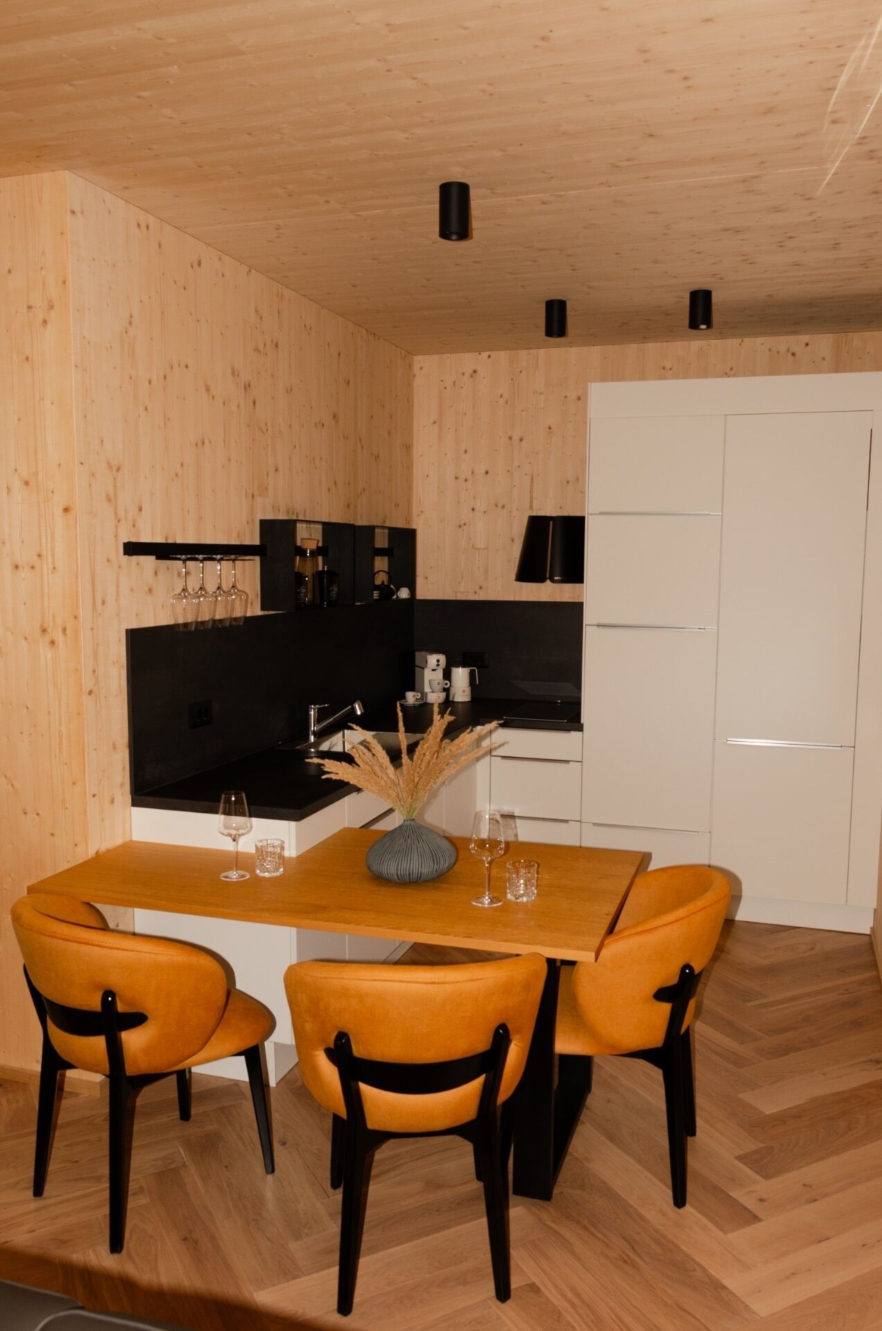 A modern kitchen with wooden walls, a black countertop, and a white refrigerator. There is a dining table with two chairs and a vase with dried plants.