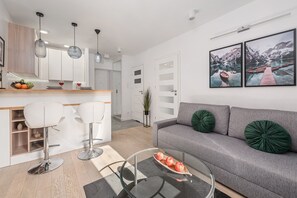 Een moderne woonkamer met een grijze bank, twee witte barkrukken, een glazen salontafel en een houten dressoir.