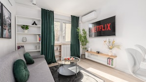 Un salon moderne avec un canapé gris, une table basse en verre, une bibliothèque, un téléviseur et un radiateur.