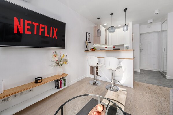 Een moderne woonkamer met een grote televisie waarop 'NETFLIX' wordt weergegeven, een houten plank met boeken, een glazen salontafel en een keukenhoek met witte barkrukken.