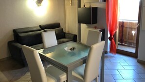Ein Wohnzimmer mit einem Glastisch, weißen Stühlen, einem dunklen Sofa, einem Fernseher auf einem Schrank und einem Balkon mit roten Vorhängen.