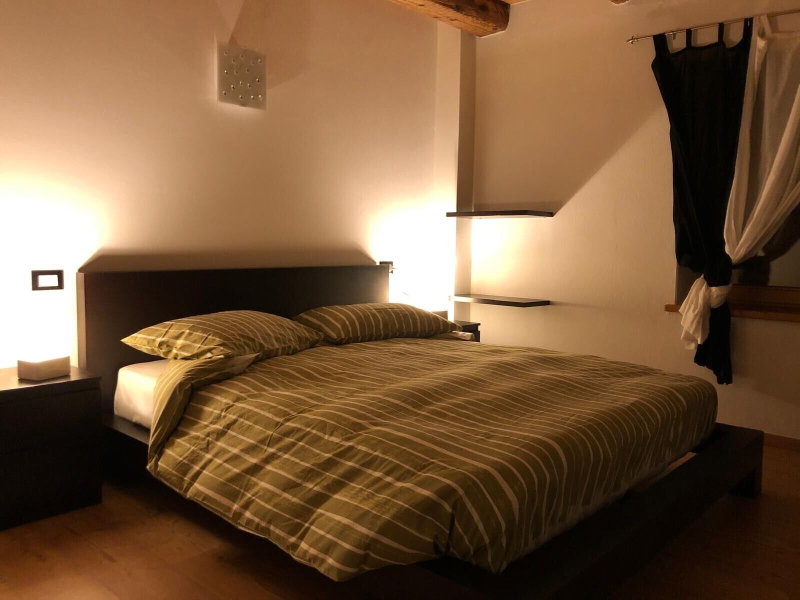 Un lit avec une couvre-lit à rayures, une table de chevet, une étagère fixée au mur et un rideau.