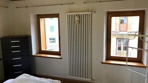 Ein Schlafzimmer mit Bett, Kommode, zwei Fenstern, Heizkörper und Balkonblick.