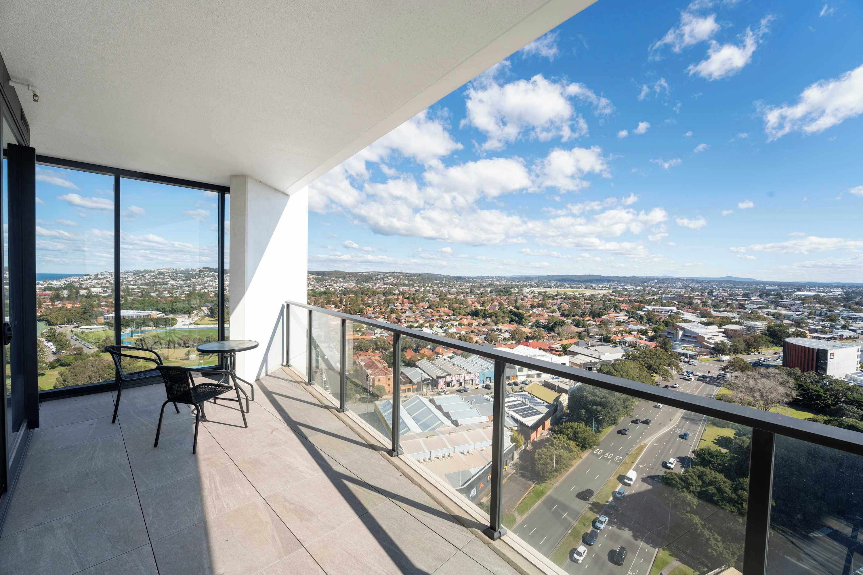 A balcony with a glass railing, a table, and two chairs, overlooking a cityscape with buildings and roads.