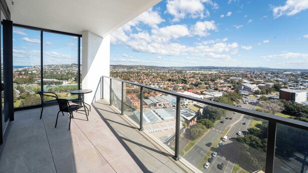 A balcony with a glass railing, a table, and two chairs, overlooking a cityscape with buildings and roads. - Prestige 2BR Stay w/ Ocean Views, Rooftop & Cinema (Newcastle West)