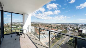A balcony with a glass railing, a table, and two chairs, overlooking a cityscape with buildings and roads.