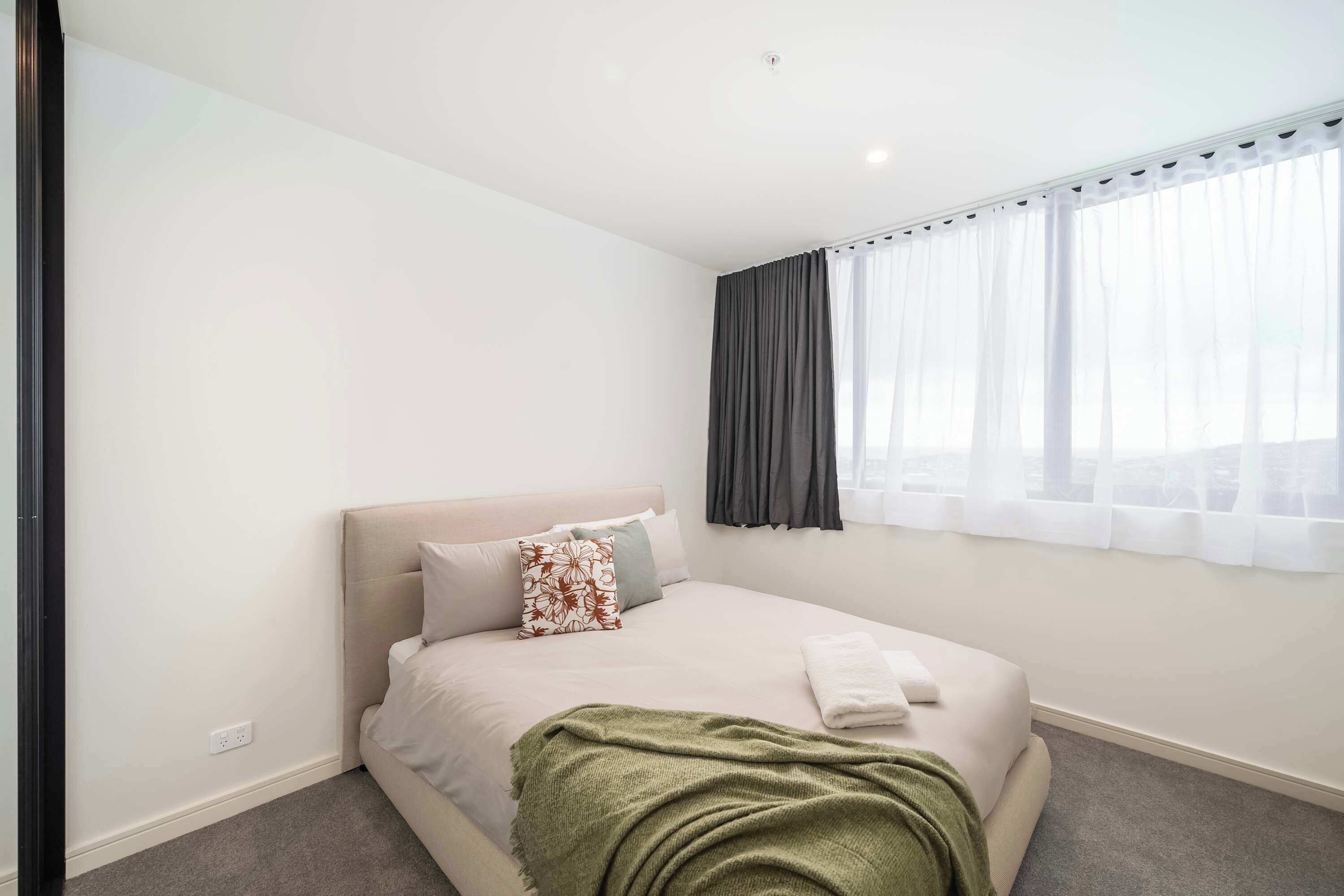 A hotel room with a bed, a window with curtains, a power outlet, and a green throw blanket.