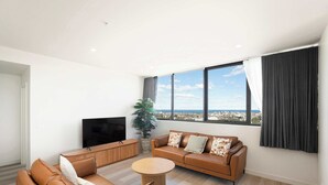 A modern living room with a sofa set, a coffee table, a TV, and a large window with a city view. - Prestige 2BR Stay w/ Ocean Views, Rooftop & Cinema (Newcastle West)