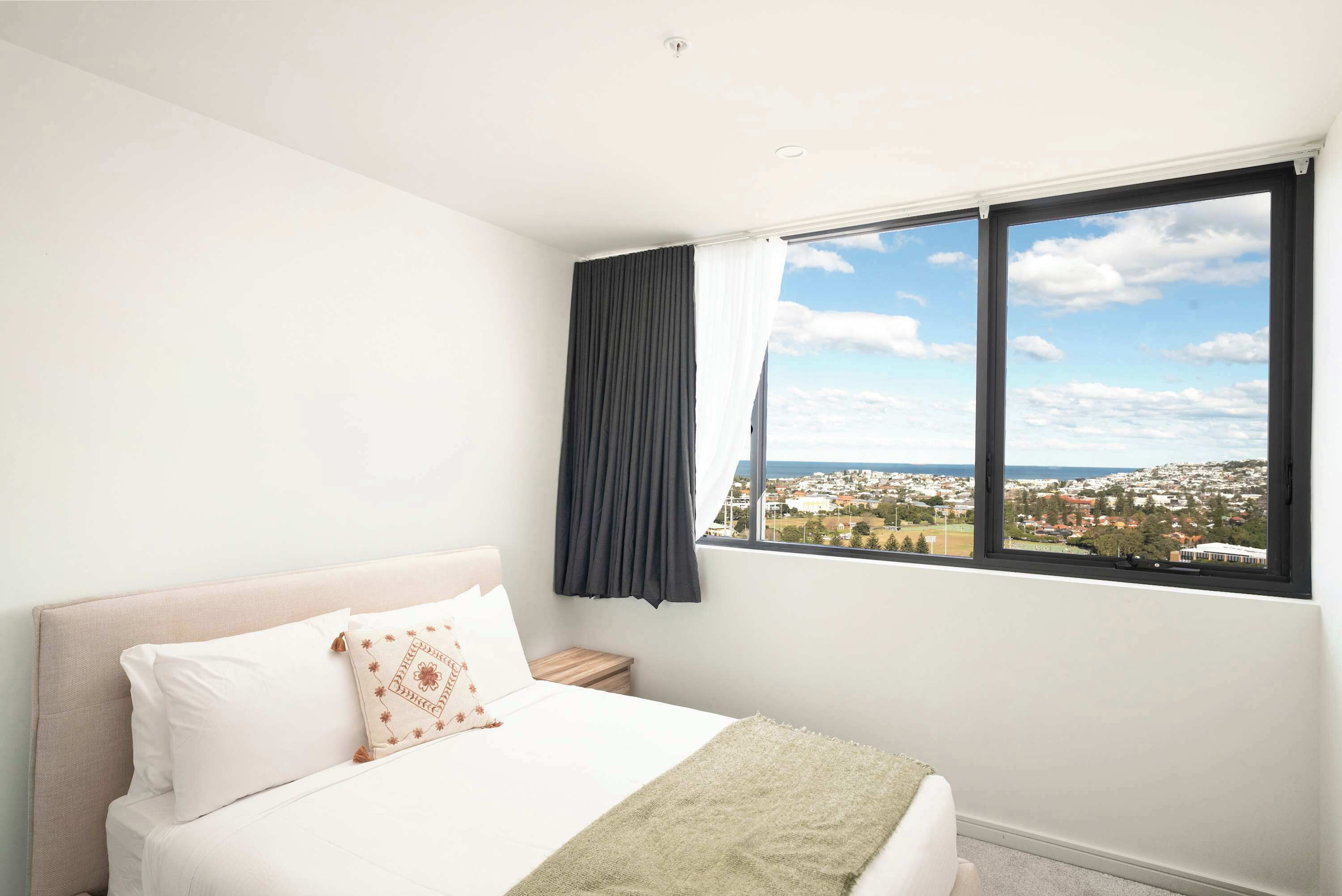 A bedroom with a bed, a nightstand, a window with a view of a town and sea, and a curtain.