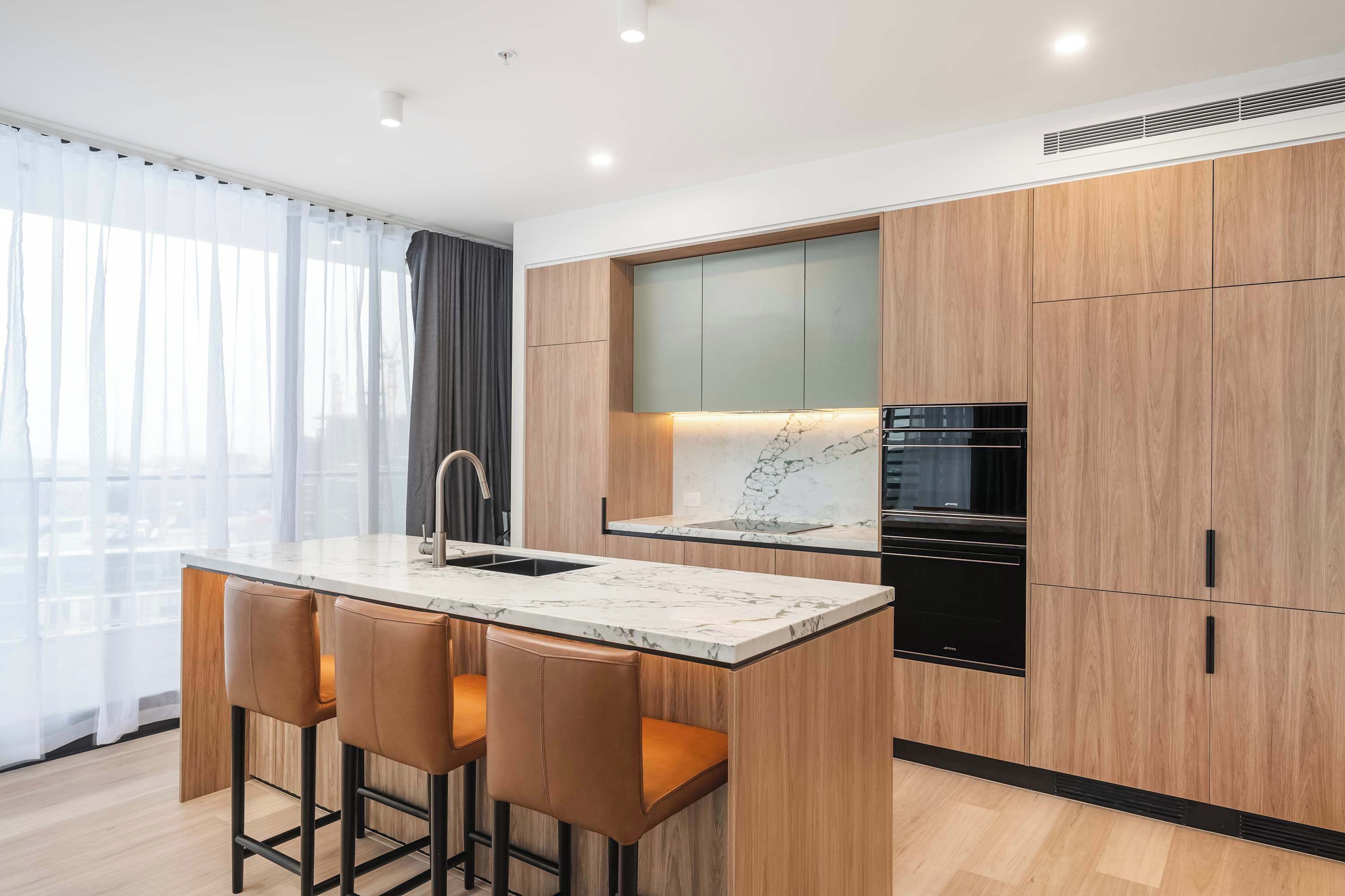 A modern kitchen with a marble island, bar stools, wooden cabinets, and a black dishwasher.