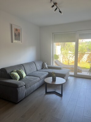 A modern living room with a grey sofa, a small round table with a plant, a framed picture on the wall, and a sliding glass door leading to an outdoor area. - Apartment Mariner 2 With Heated Pool (Nin)