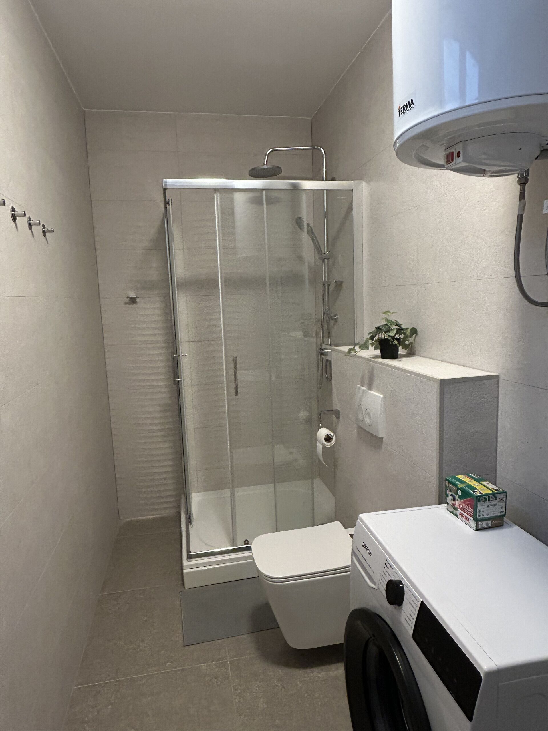 Een badkamer met een douchecabine van glas, een toilet, een wasmachine en een kleine potplant.