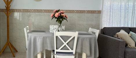 A dining area with a table set for two, a white chair, a gray sofa, and a decorative vase with flowers.