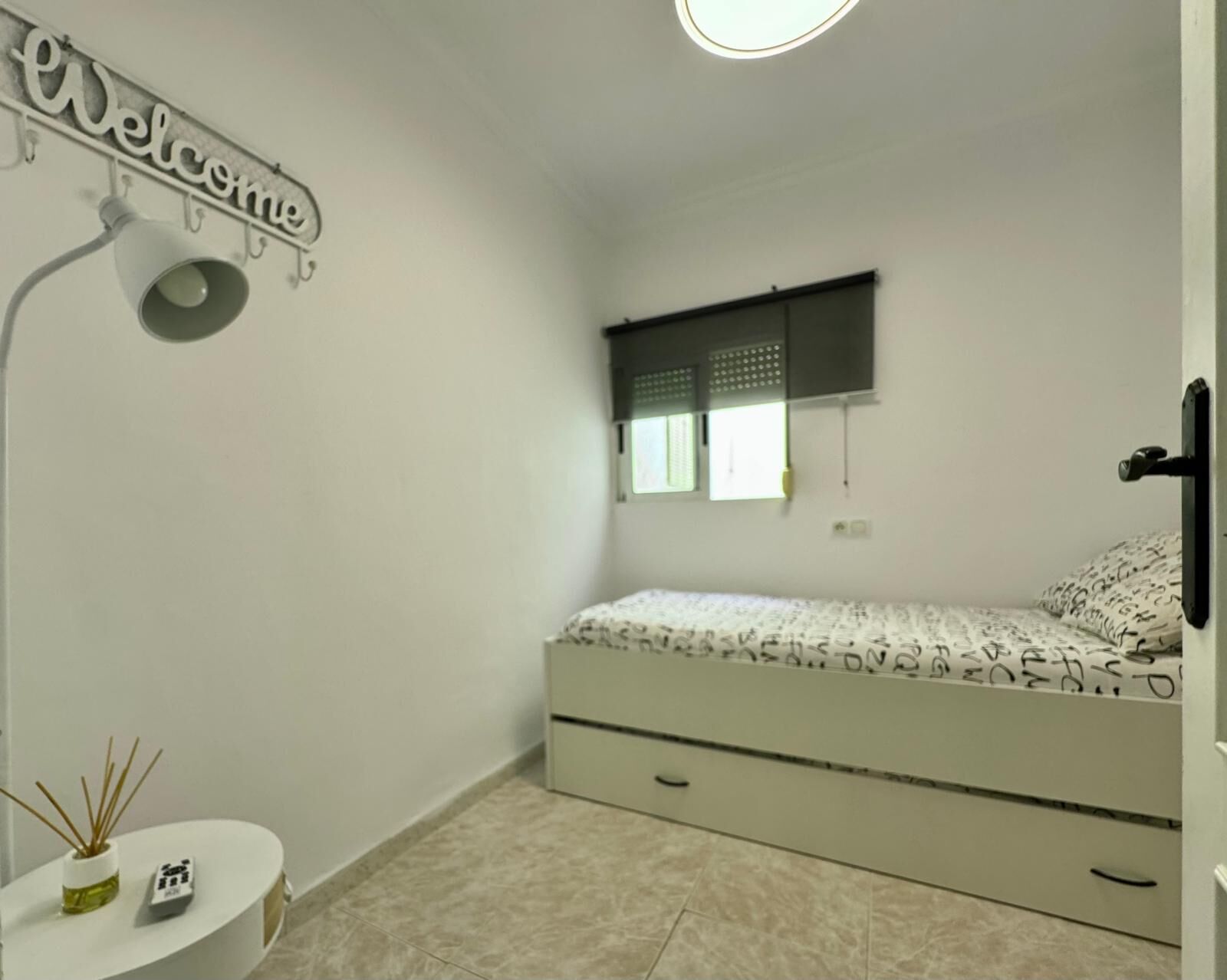 A bedroom with a bed, a bedside table with a lamp, a 'Welcome' sign, a window with blinds, and a door.