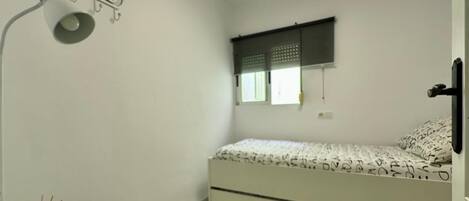A bedroom with a bed, a bedside table with a lamp, a 'Welcome' sign, a window with blinds, and a door.