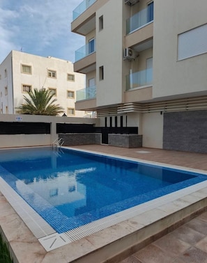 Une piscine dont le bord est revêtu de carrelage, entourée d’un petit muret et bordée d’un bâtiment doté de balcons.