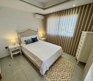 Une chambre à coucher comprenant un lit, des tables de chevet, un appareil de climatisation, une armoire et une fenêtre avec des rideaux.