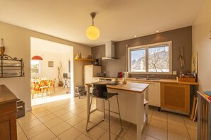 Eine moderne Küche mit einer zentralen Kochinsel, einem Essbereich mit Tisch und Stühlen sowie einem großen Fenster, das viel Tageslicht hereinlässt.
