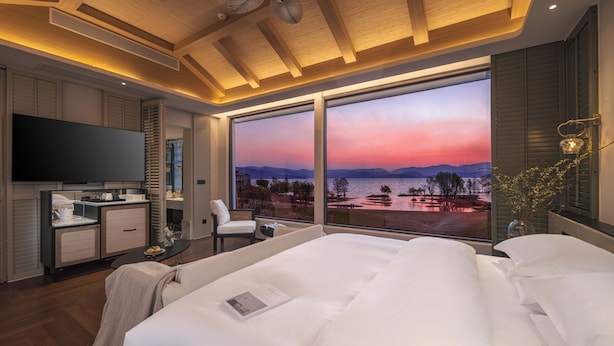 Ein modernes Schlafzimmer mit einem großen Bett, einem Flachbildfernseher, einem Schreibtisch und Blick auf einen Sonnenuntergang über dem Wasser.