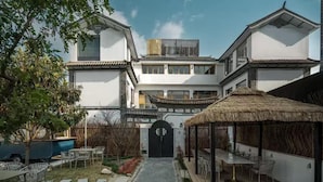Ein traditionelles Gebäude mit einem zentralen Eingang, einer überdachten Außenbereich-Sitzgelegenheit und einem kleinen Garten.