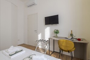 Habitación de hotel con una cama, un escritorio con una silla, un televisor, una mesita con un jarrón y una botella, y una ventana.