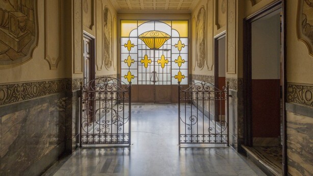 Ein Flur mit Marmorboden, aufwendigen Wandverzierungen und einem großen Fenster mit gelb-weißen Mustern.