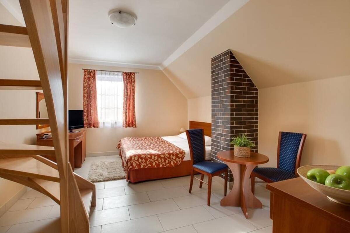 A hotel room with a bed, a desk, a chair, a small table, a plant, and a bowl of fruit.