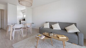 Una sala de estar moderna con un sofá gris, cojines blancos, una mesa de centro de madera y un comedor con una mesa y sillas blancas.