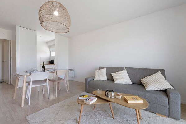 Un salon moderne avec un canapé gris, des coussins blancs, une table basse en bois et un coin repas avec une table et des chaises blanches.