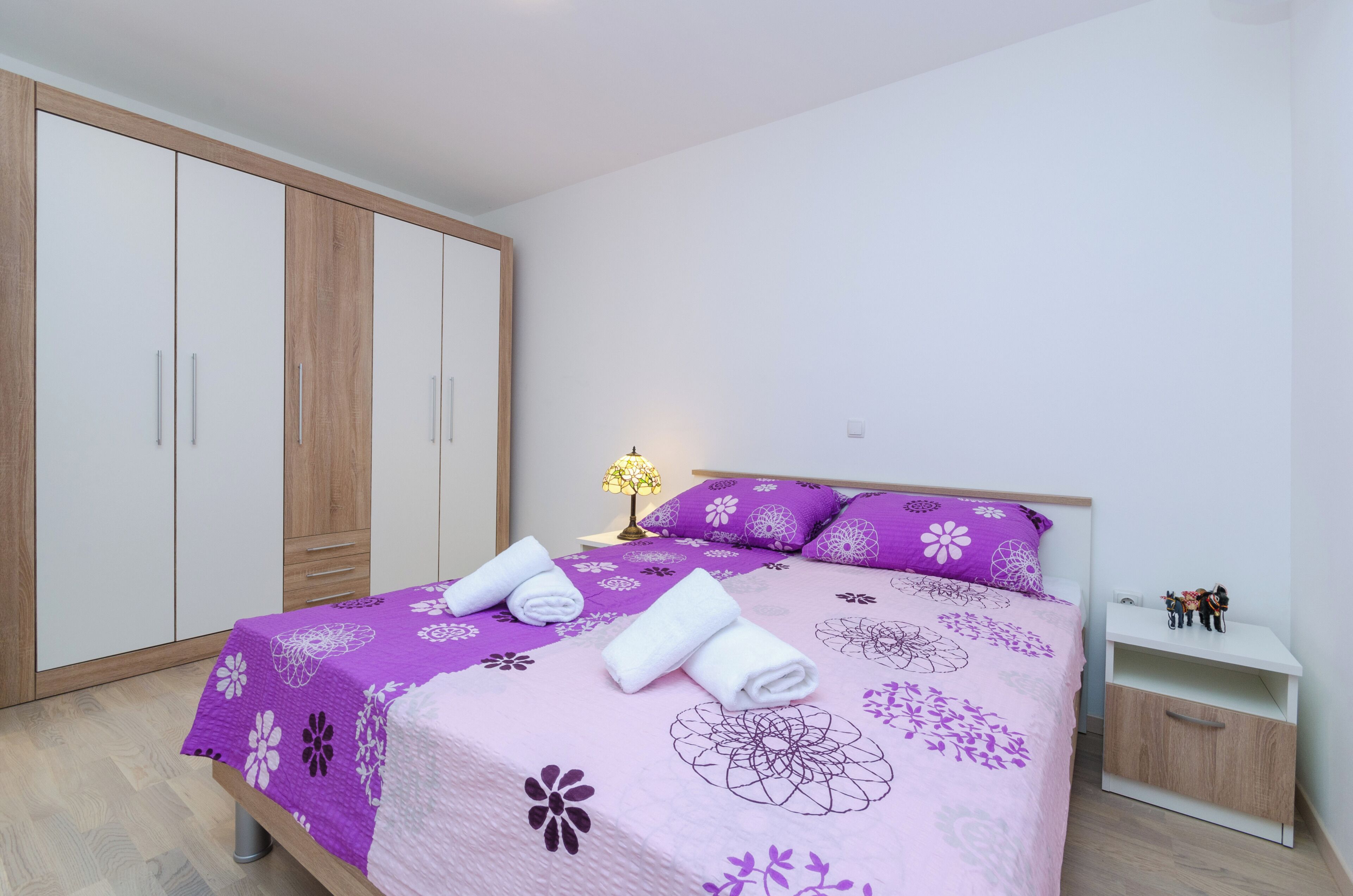 A bedroom with a bed, a nightstand, a lamp, a wardrobe, and a small shelf.
