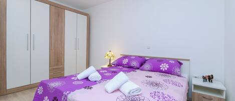 A bedroom with a bed, a nightstand, a lamp, a wardrobe, and a small shelf.