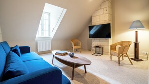 Un salon moderne avec un canapé bleu, une table basse en bois, des fauteuils en osier, un téléviseur à écran plat fixé à un mur en pierre et un lampadaire.