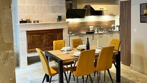 Un coin repas avec une table en bois et des chaises jaunes, une cheminée en pierre et une cuisine équipée d’appareils électroménagers modernes.