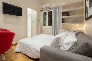 A bedroom with a bed, a grey sofa, a red chair, a TV, a window with curtains, a bookshelf, and an air conditioning unit. - Marasli 29 1bdr·Serenity Escape with Jacuzzi (Athens)