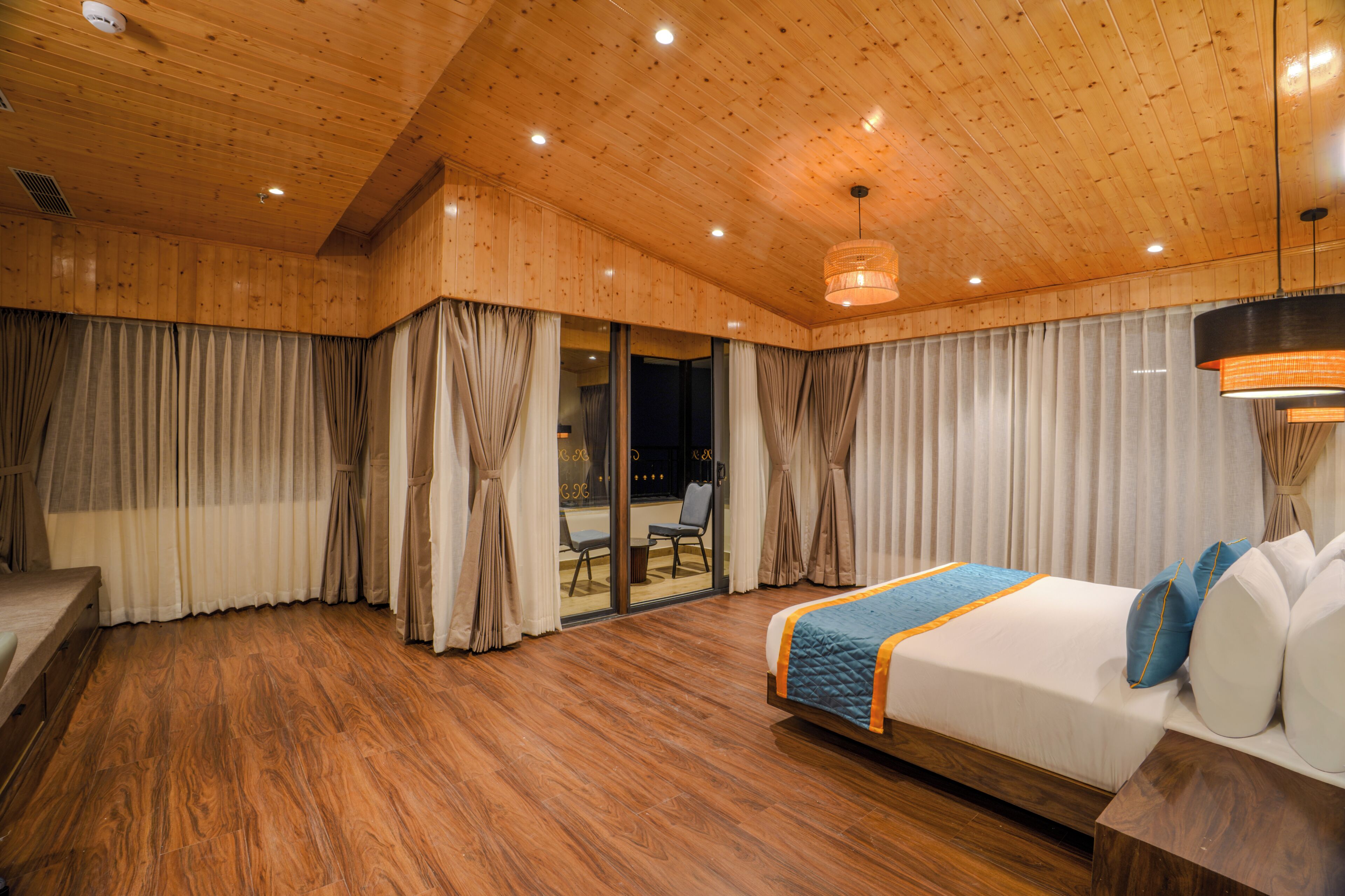 Habitación de hotel con techo de madera, cama con colcha azul y vistas al exterior a través de un ventanal.