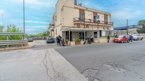 Un edificio con balcón, un coche estacionado adelante y una persona junto a la entrada.