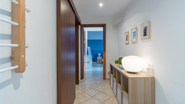 Un pasillo con una puerta de madera, un toallero blanco, una lámpara de pared y una pequeña mesa con una lámpara y una botella.