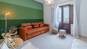 Una habitación con pared verde, sofá marrón, una mesita redonda pequeña, una silla blanca, una lámpara colgante y vistas a través de una ventana con cortinas.