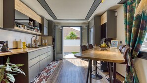 Een moderne keuken met inbouwapparatuur, een zithoek met een houten tafel en stoelen, en uitzicht door een open deur op een buitenruimte.